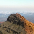 A breathtaking view from Kalsubai Peak, Maharashtra’s highest point, showing a mist-covered trail, lush green hills, and dramatic clouds rolling over the mountain landscape.