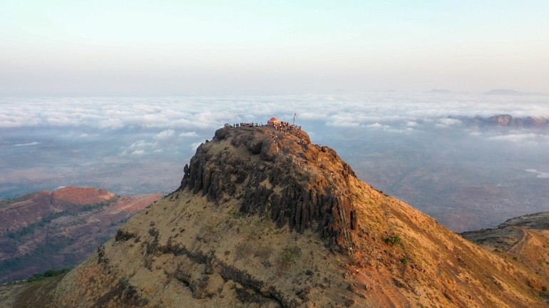 A breathtaking view from Kalsubai Peak, Maharashtra’s highest point, showing a mist-covered trail, lush green hills, and dramatic clouds rolling over the mountain landscape.