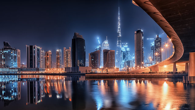 Nighttime view of Business Bay in Dubai, showcasing illuminated skyscrapers, modern architecture, and vibrant city lights reflecting on the water.