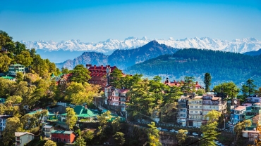 Panoramic cityscape of Shimla, Himachal Pradesh, featuring colonial-era buildings, lush green hills, and mist-covered mountain views under a soft blue sky.