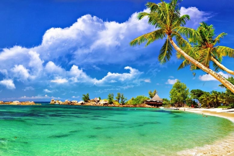 A panoramic view of the coastal city of Port Blair featuring turquoise waters, green hills, and scattered buildings along the shoreline under a partly cloudy sky.