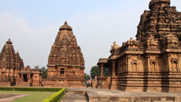 Historic Chalukyan temple complex made of intricately carved reddish-brown stone, with tall spires and sculptures, set against a dramatic cloudy sky in Pattadakal, Karnataka.