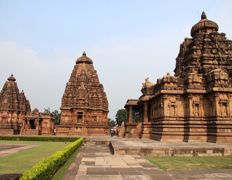 Historic Chalukyan temple complex made of intricately carved reddish-brown stone, with tall spires and sculptures, set against a dramatic cloudy sky in Pattadakal, Karnataka.