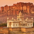 A vibrant cityscape of Jodhpur, Rajasthan, featuring blue-painted houses densely packed around the base of the historic Mehrangarh Fort under a clear sky.