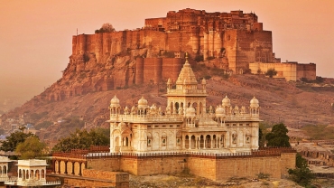 A vibrant cityscape of Jodhpur, Rajasthan, featuring blue-painted houses densely packed around the base of the historic Mehrangarh Fort under a clear sky.