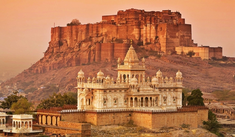 A vibrant cityscape of Jodhpur, Rajasthan, featuring blue-painted houses densely packed around the base of the historic Mehrangarh Fort under a clear sky.