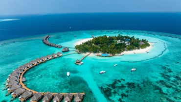 A breathtaking aerial view of a Maldives island featuring white sandy beaches, overwater villas extending into the turquoise ocean, and lush greenery surrounded by vibrant coral reefs under a clear blue sky.