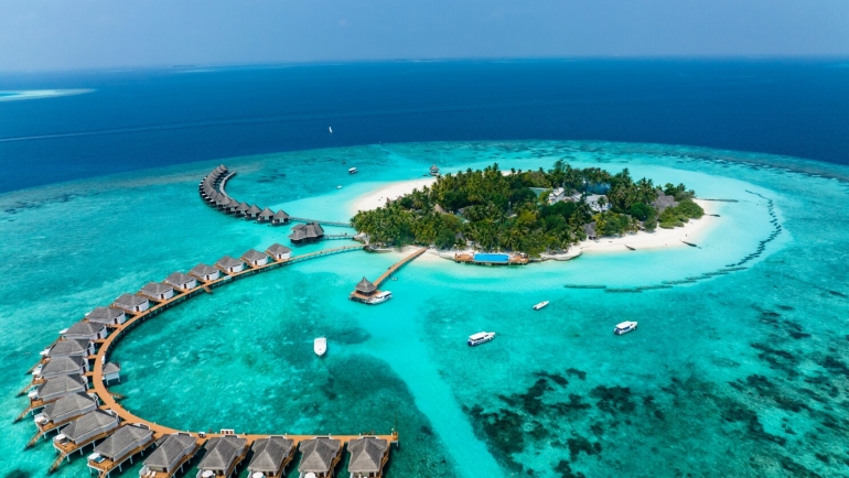 A breathtaking aerial view of a Maldives island featuring white sandy beaches, overwater villas extending into the turquoise ocean, and lush greenery surrounded by vibrant coral reefs under a clear blue sky.