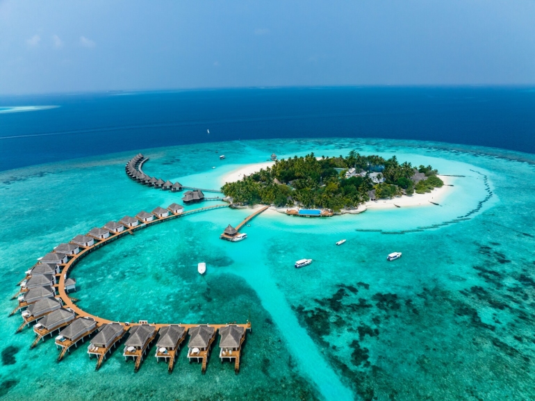 A breathtaking aerial view of a Maldives island featuring white sandy beaches, overwater villas extending into the turquoise ocean, and lush greenery surrounded by vibrant coral reefs under a clear blue sky.