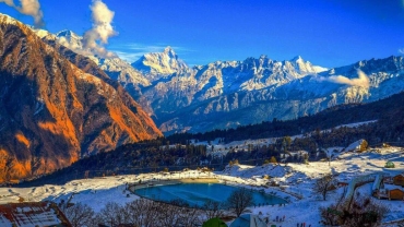 A scenic winter landscape of Auli, India, showing snow-covered slopes, pine trees, and a ski resort area under a clear blue sky, perfect for snowfall and winter sports.
