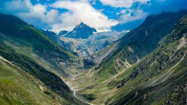 A scenic view of Hampta Pass with a narrow mountain trail winding through green meadows, surrounded by rugged snow-capped peaks and dramatic rocky terrain under a partly cloudy sky.