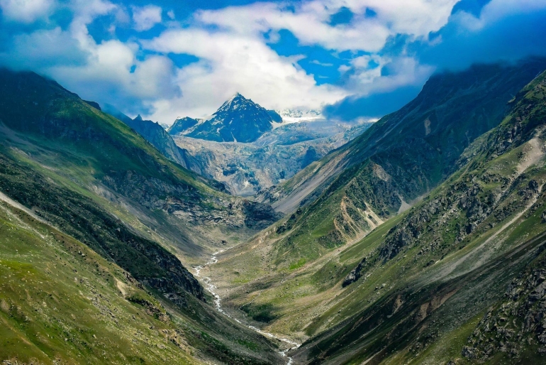 A scenic view of Hampta Pass with a narrow mountain trail winding through green meadows, surrounded by rugged snow-capped peaks and dramatic rocky terrain under a partly cloudy sky.