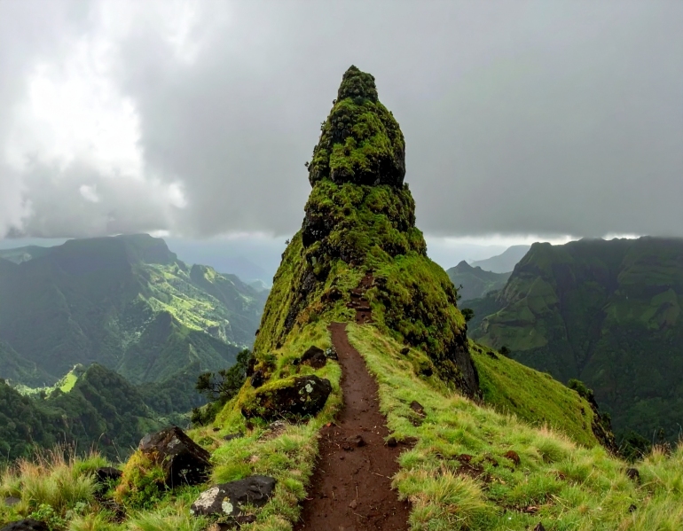 A narrow rocky pinnacle at Irshalgad Fort in Maharashtra, surrounded by lush green grass and misty Western Ghats under a cloudy monsoon sky, with a dirt trail leading toward the peak.