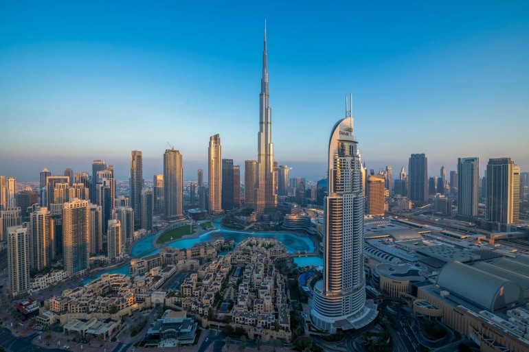 A majestic view of the Burj Khalifa in Dubai, soaring into a clear blue sky, surrounded by modern skyscrapers and urban landscape in the foreground.