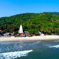 Aerial view of Ganpatipule Beach in Ratnagiri, featuring the iconic Ganpatipule Temple with a white spire and red base, set against a lush green hill, with waves gently hitting the sandy shoreline under a clear blue sky