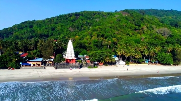 Aerial view of Ganpatipule Beach in Ratnagiri, featuring the iconic Ganpatipule Temple with a white spire and red base, set against a lush green hill, with waves gently hitting the sandy shoreline under a clear blue sky