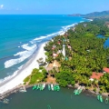 Aerial view of a tropical beach town with palm trees, red-roofed buildings, and green fishing boats docked along a river meeting the Arabian Sea. Gentle waves roll onto the white sandy beach under a clear blue sky, with a prominent white watchtower at the center of the coastal area