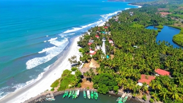 Aerial view of a tropical beach town with palm trees, red-roofed buildings, and green fishing boats docked along a river meeting the Arabian Sea. Gentle waves roll onto the white sandy beach under a clear blue sky, with a prominent white watchtower at the center of the coastal area