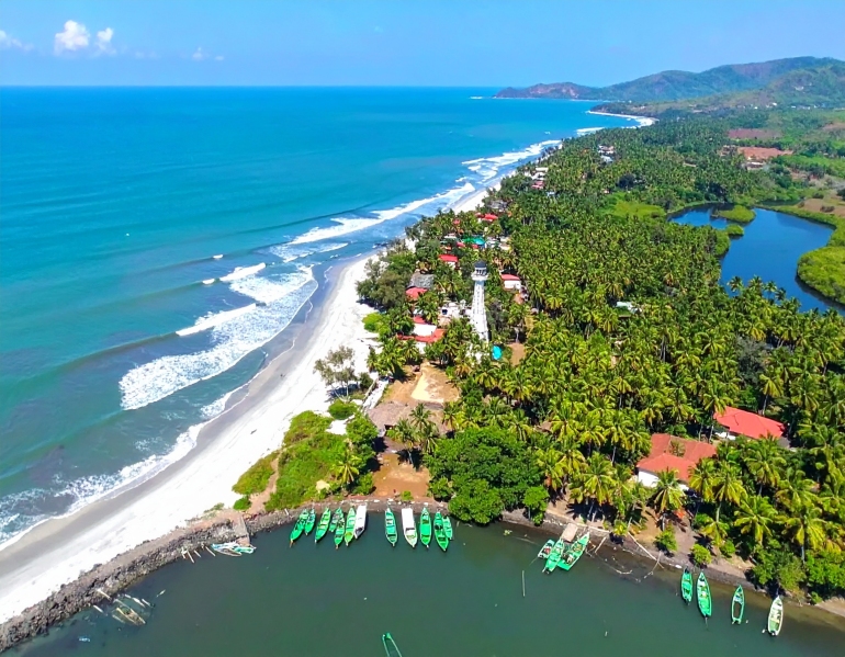 Aerial view of a tropical beach town with palm trees, red-roofed buildings, and green fishing boats docked along a river meeting the Arabian Sea. Gentle waves roll onto the white sandy beach under a clear blue sky, with a prominent white watchtower at the center of the coastal area