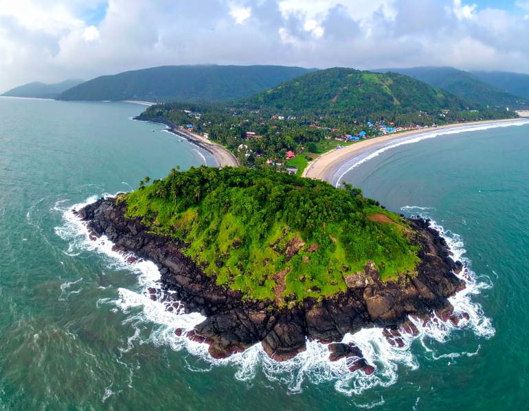 Aerial view of Harihareshwar and Diveagar coastline in Maharashtra, featuring a lush green hill surrounded by the Arabian Sea, curved beach with gentle waves, and a small seaside village nestled along the shore.