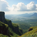 People standing on the edge of Visapur Fort, surrounded by steep green cliffs and overlooking a vast valley with distant mountains under a blue sky with scattered clouds.
