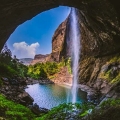 View of Devkund Waterfall cascading from a cliff into a clear pool, seen from inside a cave with lush greenery and dramatic rock formations.