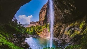 View of Devkund Waterfall cascading from a cliff into a clear pool, seen from inside a cave with lush greenery and dramatic rock formations.