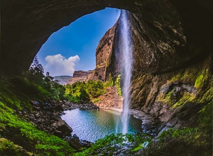 View of Devkund Waterfall cascading from a cliff into a clear pool, seen from inside a cave with lush greenery and dramatic rock formations.