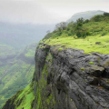 Trekker standing on a lush green cliff edge with panoramic view of the Sahyadri mountain range and valley below during monsoon.