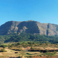 Wide view of Jivdhan Fort with Vanarlingi pinnacle standing tall on the right side under a clear blue sky.