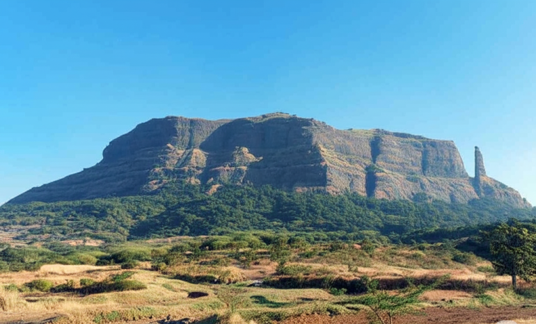 Wide view of Jivdhan Fort with Vanarlingi pinnacle standing tall on the right side under a clear blue sky.