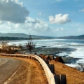 Coastal road curving along Diveagar Beach with waves crashing against the rocky shoreline under a blue sky.