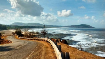 Coastal road curving along Diveagar Beach with waves crashing against the rocky shoreline under a blue sky.