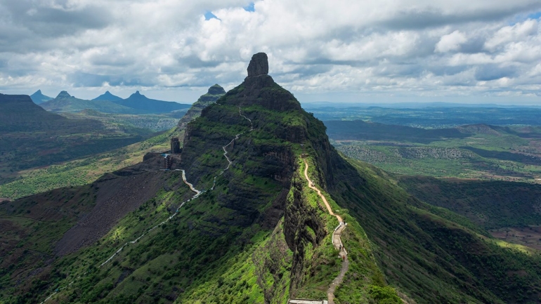 Aerial view of Mangi Tungi twin peaks with a winding path and lush green Sahyadri mountains under a cloudy sky.